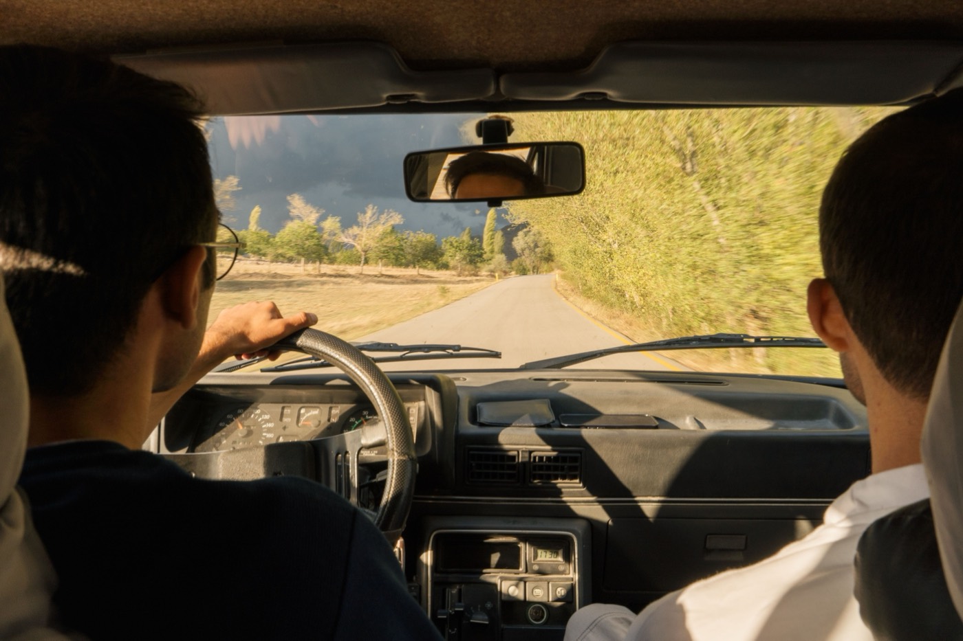 A driver and passenger on an open rural road at golden hour, seen from the back seat.